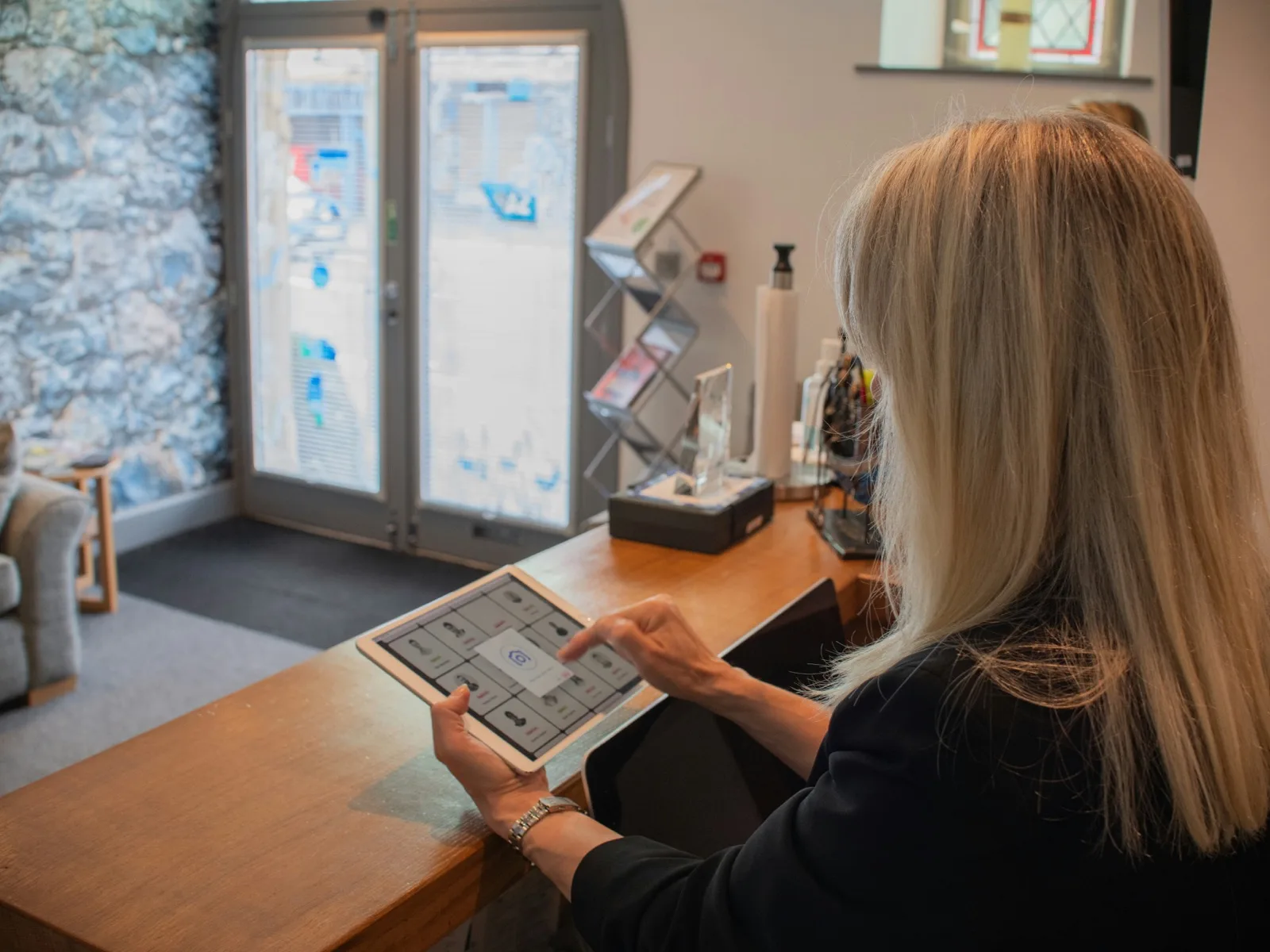A receptionist at a property's front desk working a booking grid on a tablet, the entrance and waiting area behind her — the operator side of the platform, run from the desk rather than a back office.